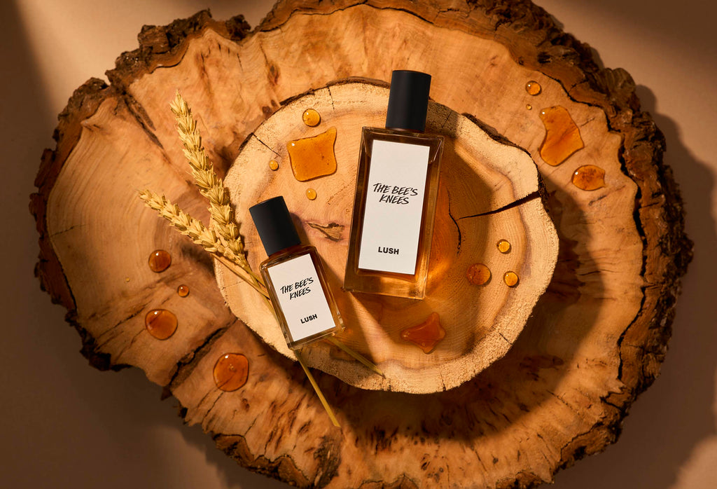 The image shows two stacked slices of wood. There is a warm light beaming over the two sizes of perfume bottles laid flat next to strands of wheat and dripped with splashes of honey.